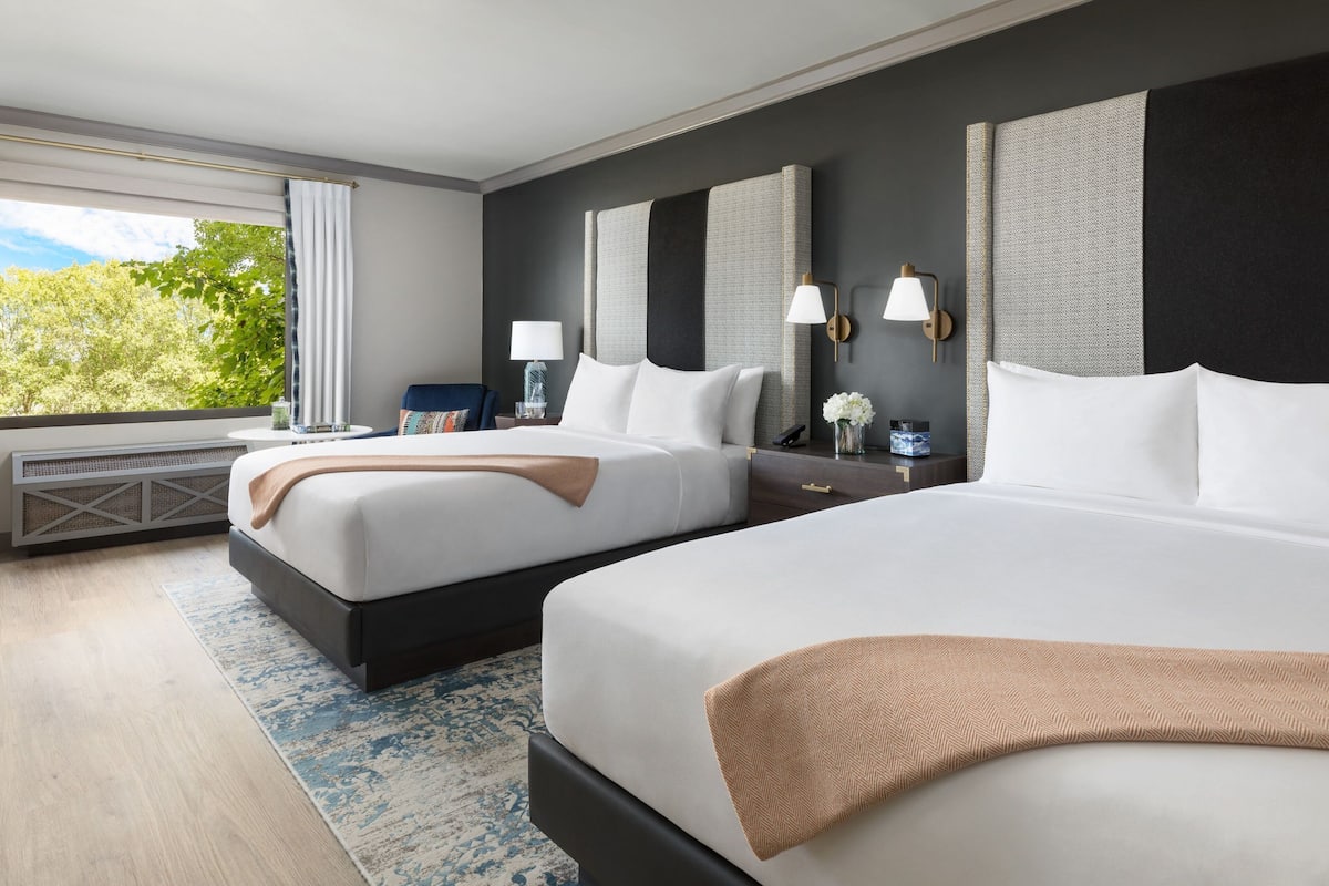 A hotel room with two beds, a large window with a view of trees, a desk with a lamp, and a nightstand with a vase of flowers.