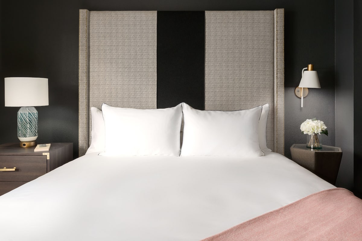 A neatly made bed with white linens, a grey textured headboard, and a bedside table with a lamp and a vase of flowers.