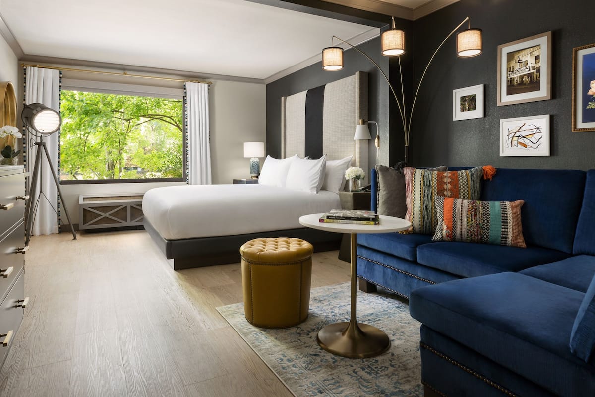 A modern hotel room with a large bed, a blue sofa, a small round table, and a view of greenery through the window.