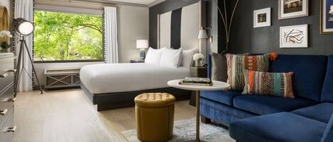 A modern hotel room with a large bed, a blue sofa, a small round table, and a view of greenery through the window.