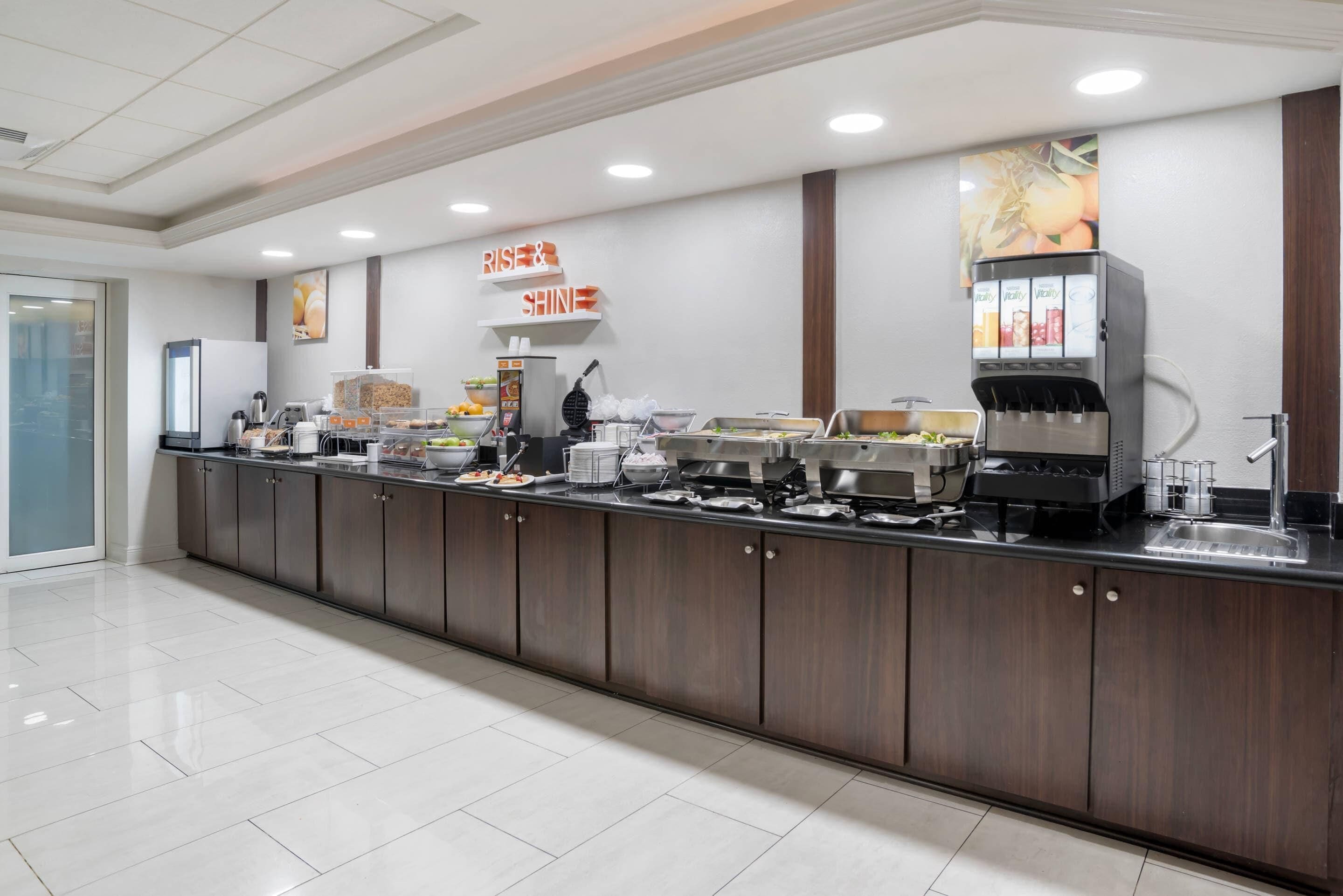A buffet setup with various food stations, a coffee machine, and a sign that reads 'RISE & SHINE'.