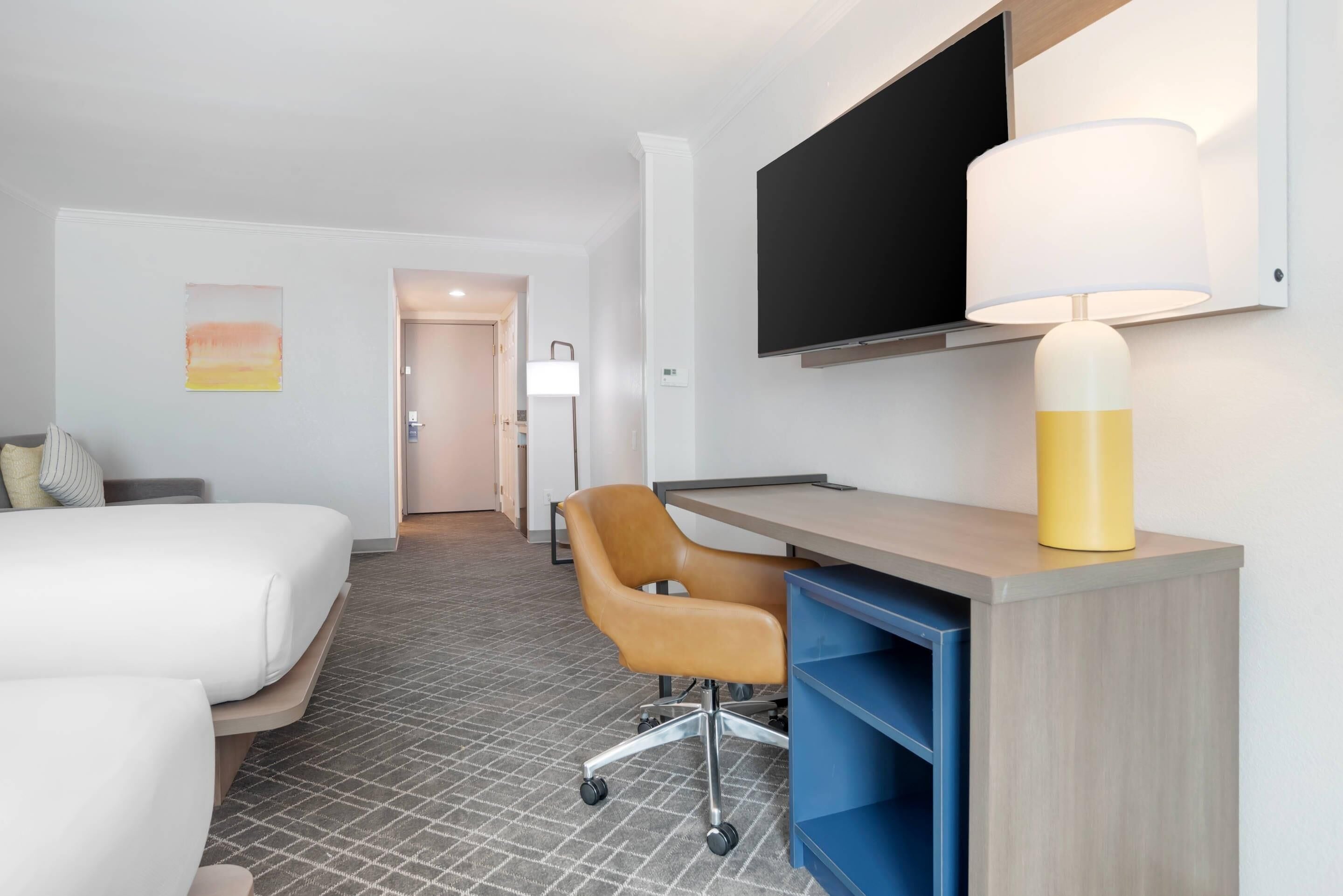 Quarto de hotel com uma cama, uma escrivaninha com cadeira, uma televisão e uma luminária.