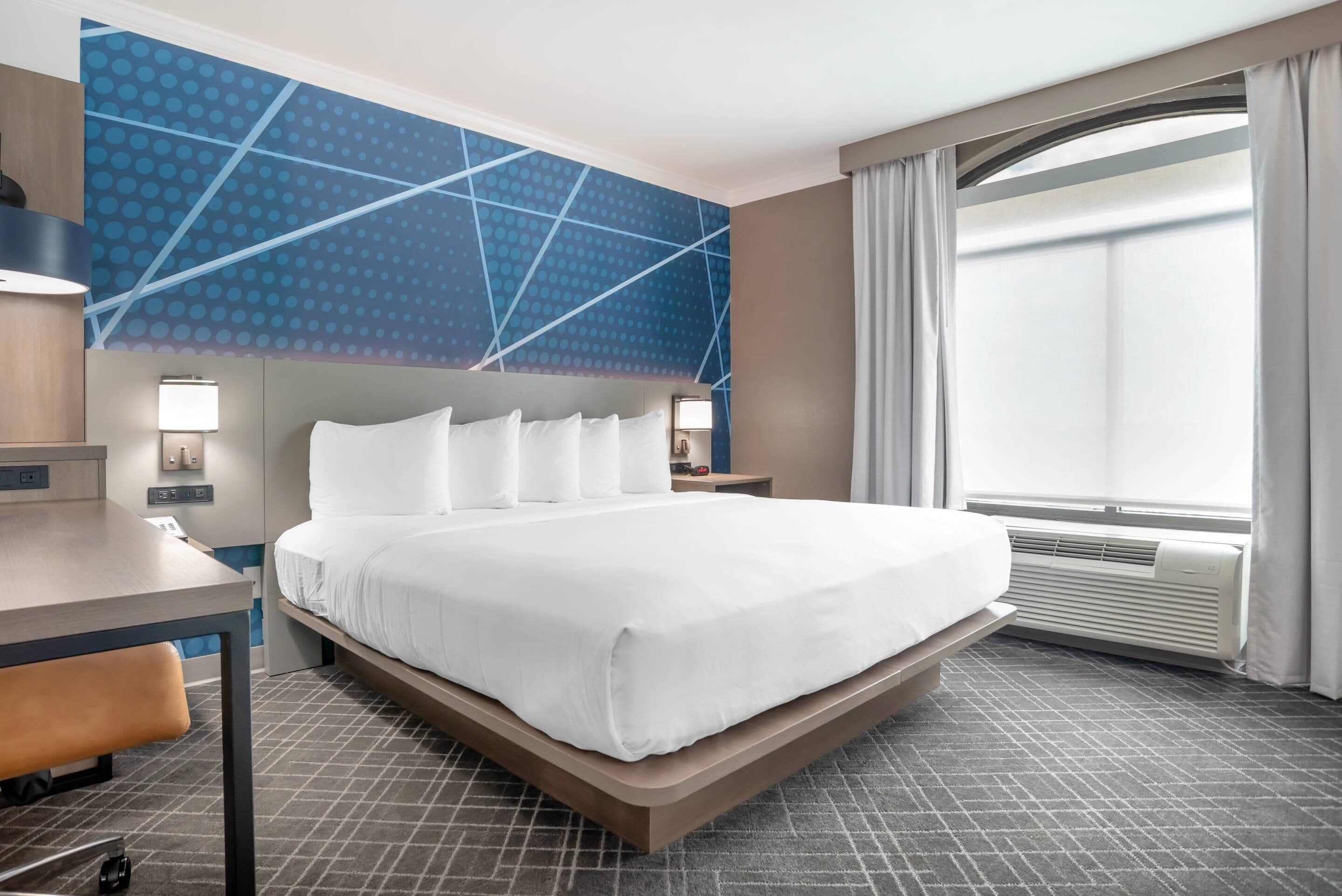A hotel room with a large bed, a desk, a chair, a nightstand, a wall-mounted lamp, and a window with curtains.