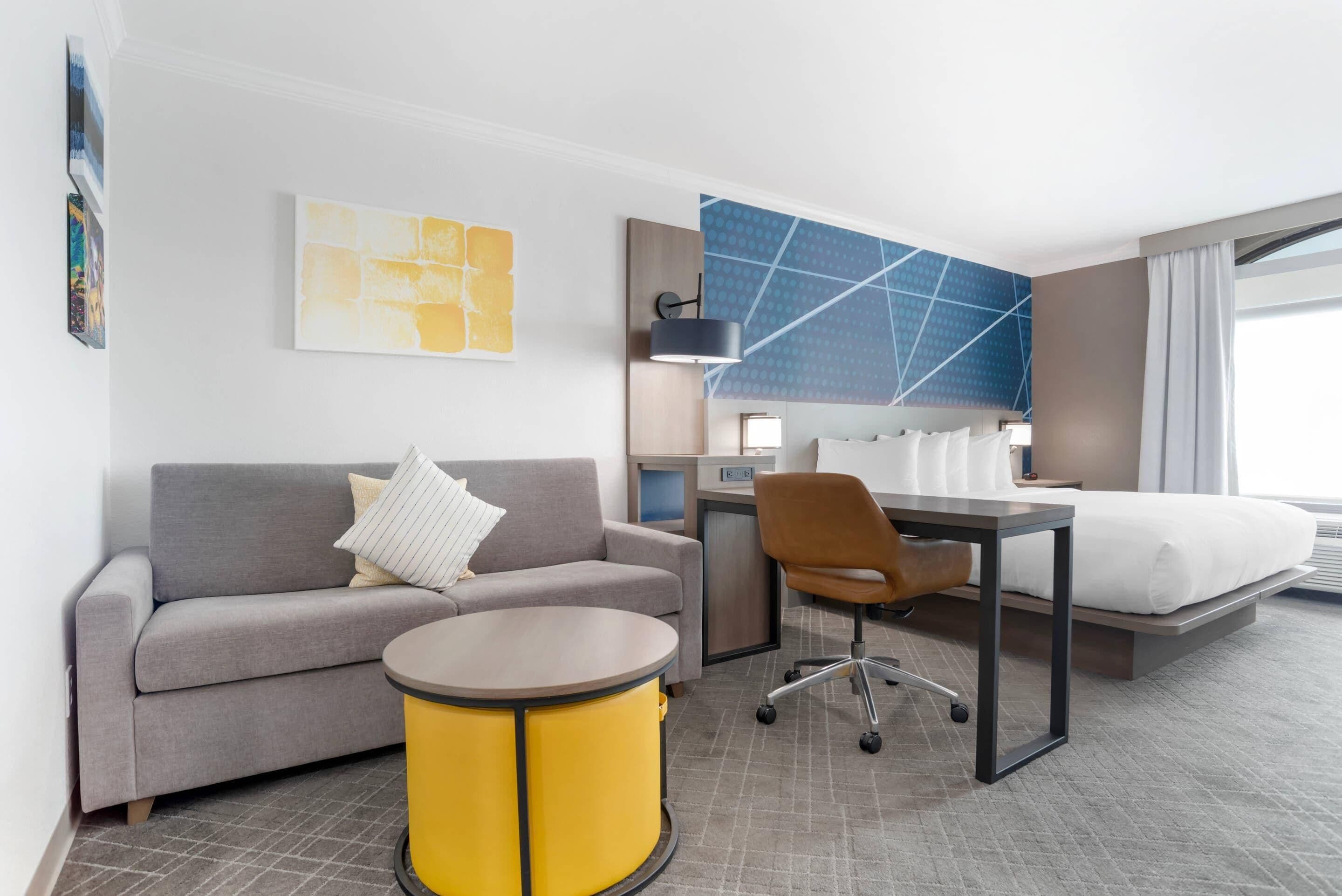 Une chambre d’hôtel avec un canapé gris, une table d’appoint jaune, un bureau avec une chaise, un lit et un grand tableau abstrait accroché au mur.