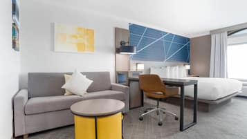 Une chambre d’hôtel avec un canapé gris, une table d’appoint jaune, un bureau avec une chaise, un lit et un grand tableau abstrait accroché au mur.