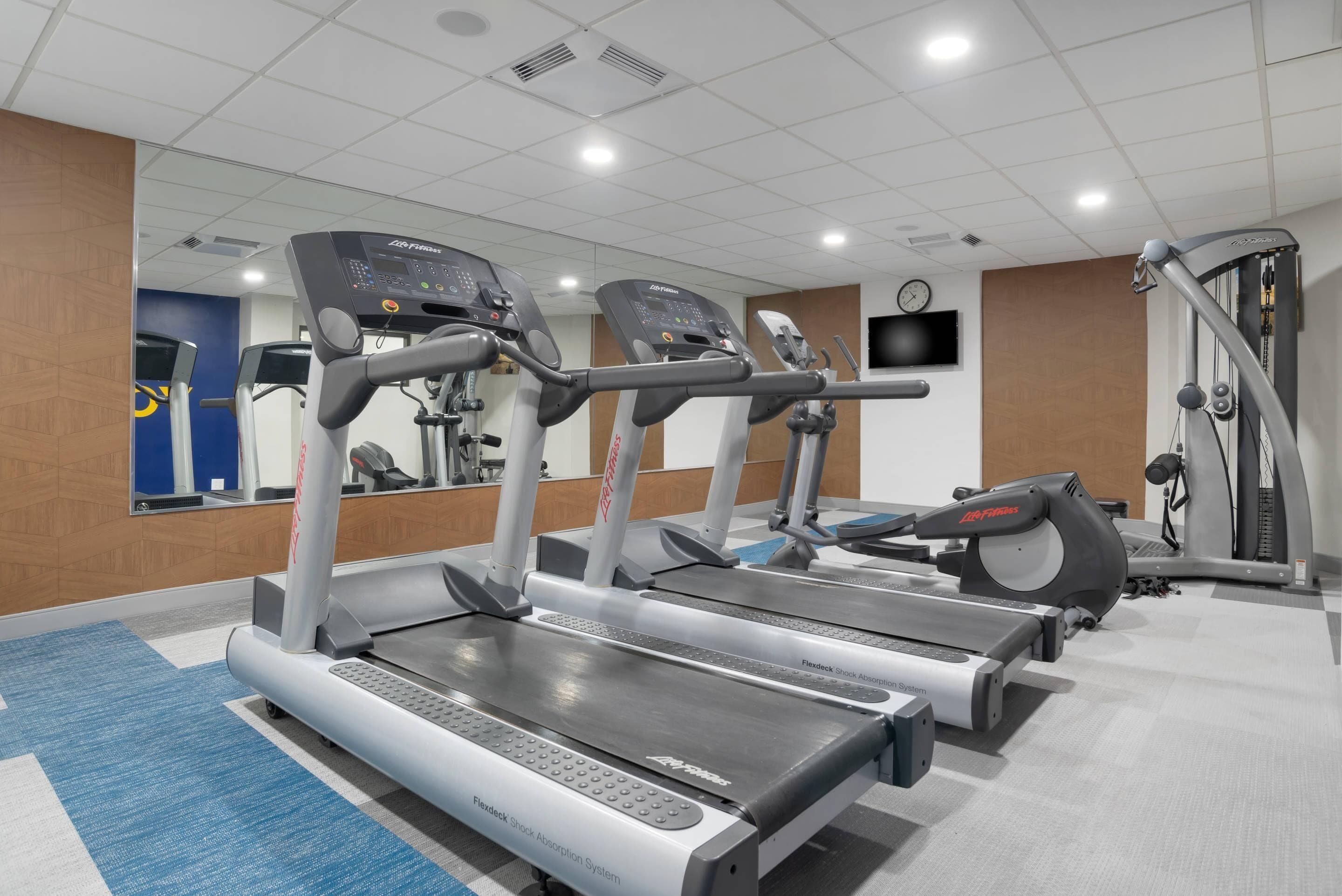 Une salle de sport équipée de tapis de course, d’un mur recouvert de miroirs et d’une horloge.