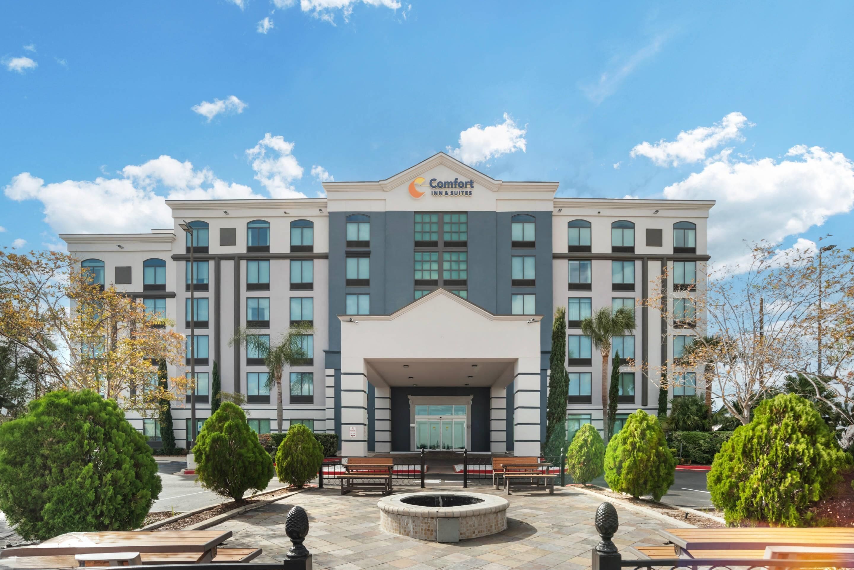 A multi-story hotel with a central entrance, surrounded by a landscaped area with benches and a circular water feature.