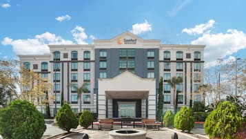 A multi-story hotel with a central entrance, surrounded by a landscaped area with benches and a circular water feature.