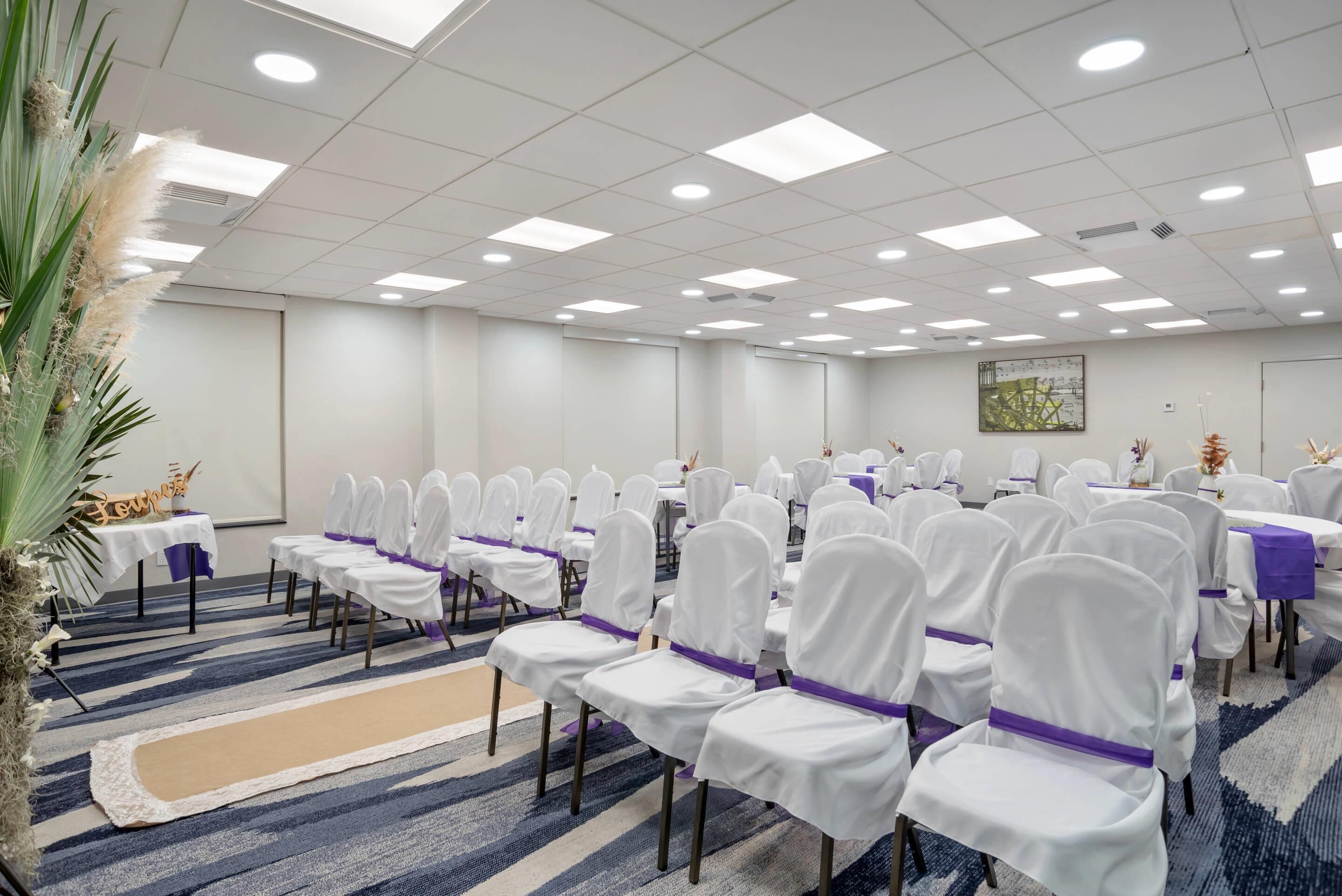 Une salle de conférence avec des rangées de chaises blanches ornées de rubans violets, des tables recouvertes de nappes blanches et un tapis à rayures bleues et blanches.