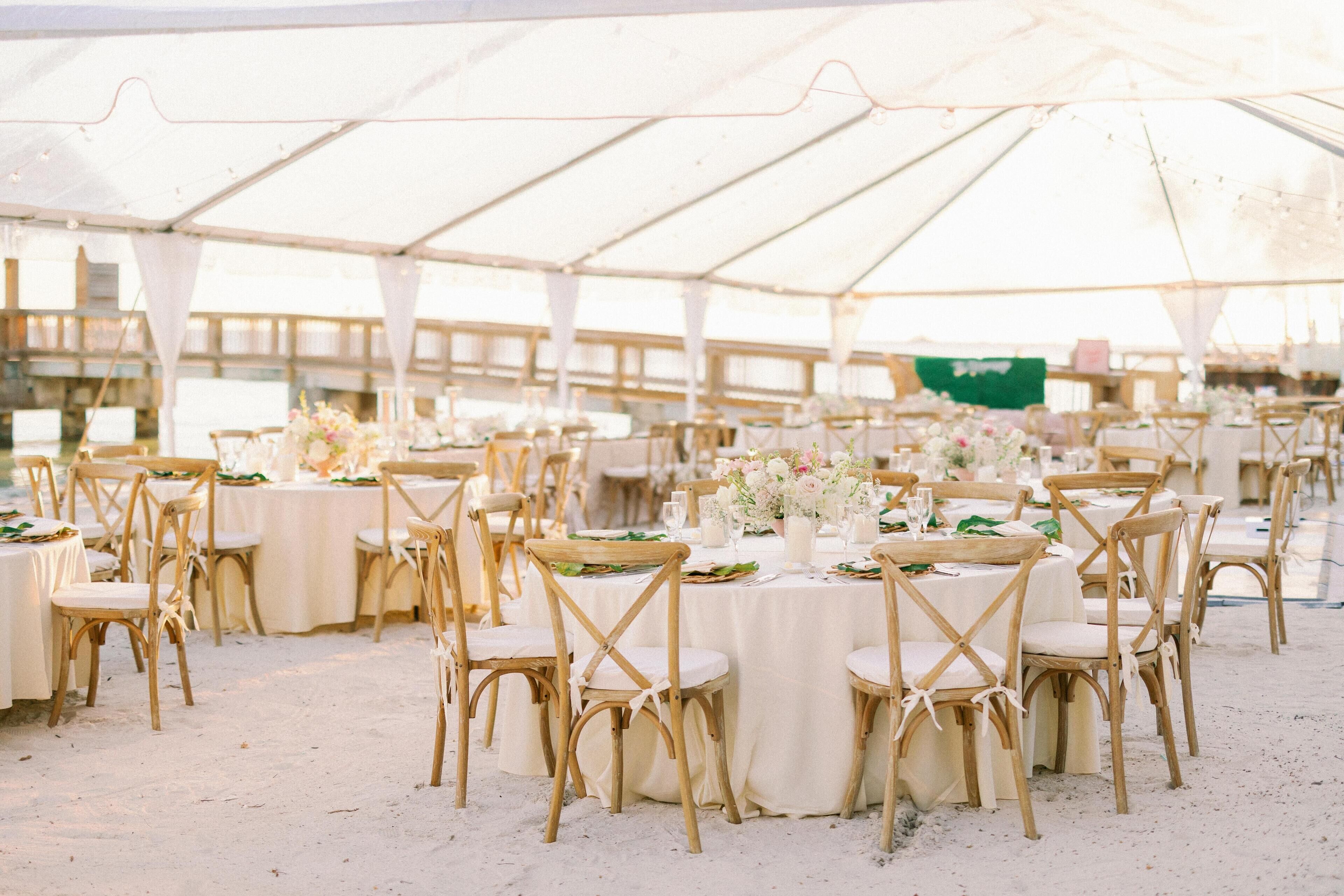A tented outdoor event space with round tables and wooden chairs, set up on a sandy ground.