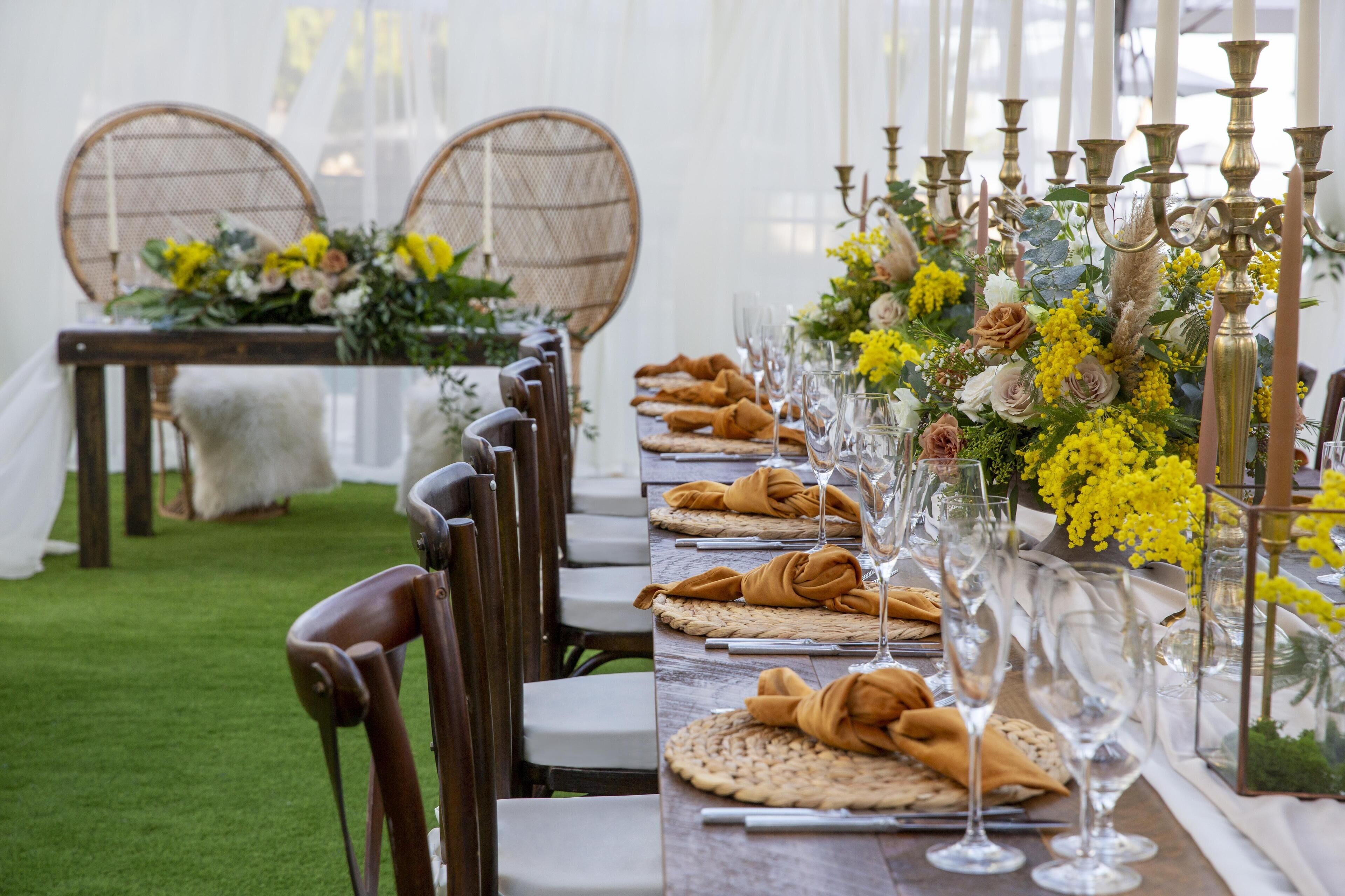 Una larga mesa de comedor con sillas, copas y un centro de mesa con flores amarillas.