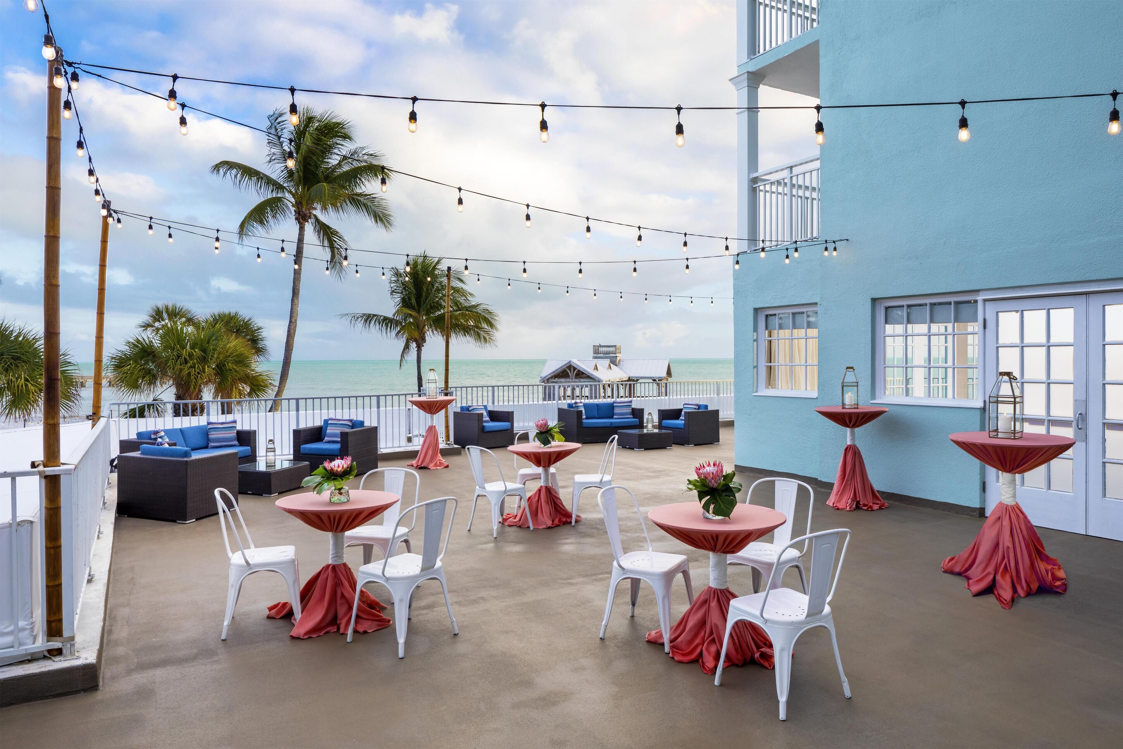 A rooftop bar with white chairs and tables, string lights, and a view of the ocean.