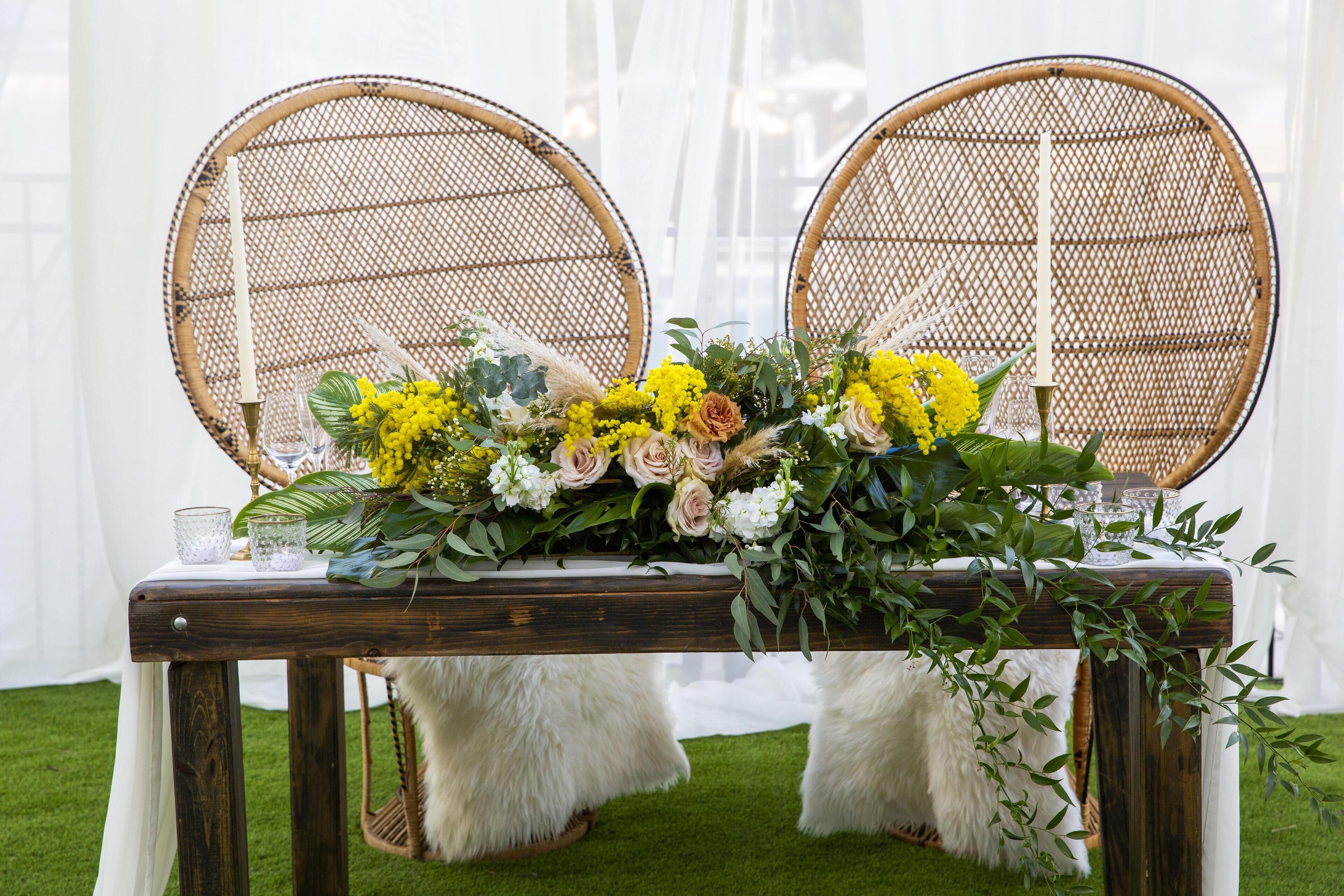 Una mesa con un centro de flores, dos sillas de mimbre y un reposapiés blanco y mullido.