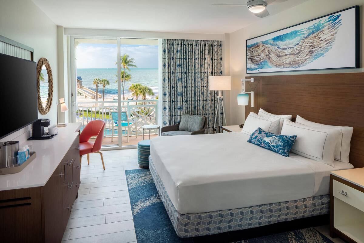 Een hotelkamer met een groot bed, een bureau met een televisie, een stoel, een lamp en uitzicht op het strand en de oceaan.