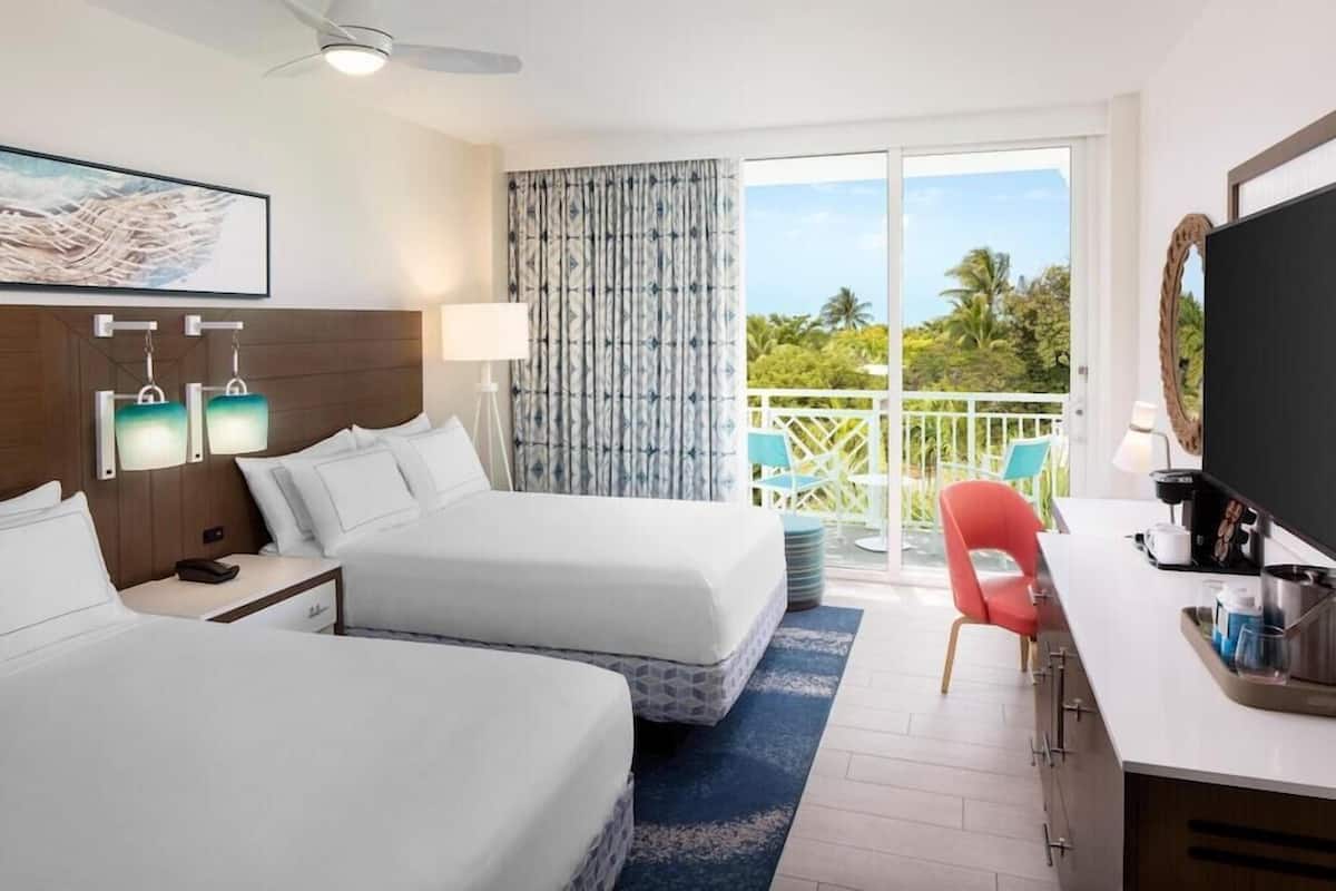 Une chambre d’hôtel avec deux lits, un bureau, une télévision et un balcon offrant une vue sur les palmiers.