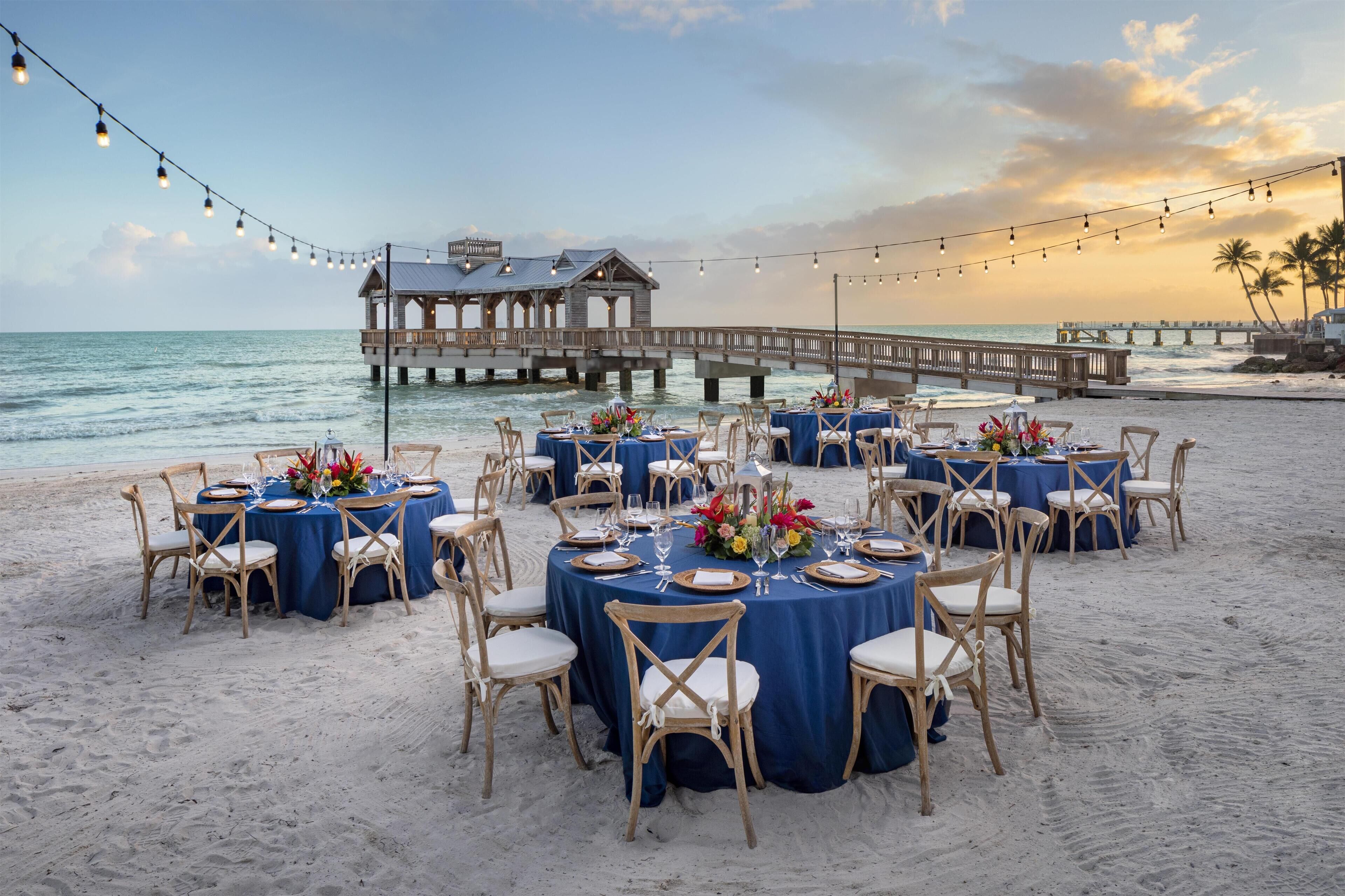 A beachside dining setup with round tables, chairs, and string lights.