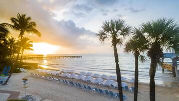 Une plage bordée de palmiers, avec des chaises longues et des parasols, sous un ciel embrasé par le coucher de soleil.
