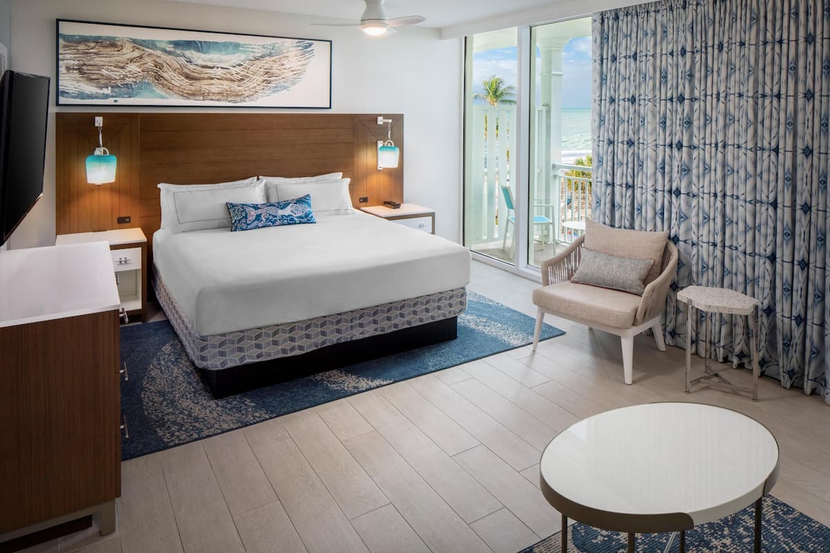 Hotelkamer met een groot bed, een stoel, een tafeltje, een televisie en een balkon met uitzicht op de oceaan.