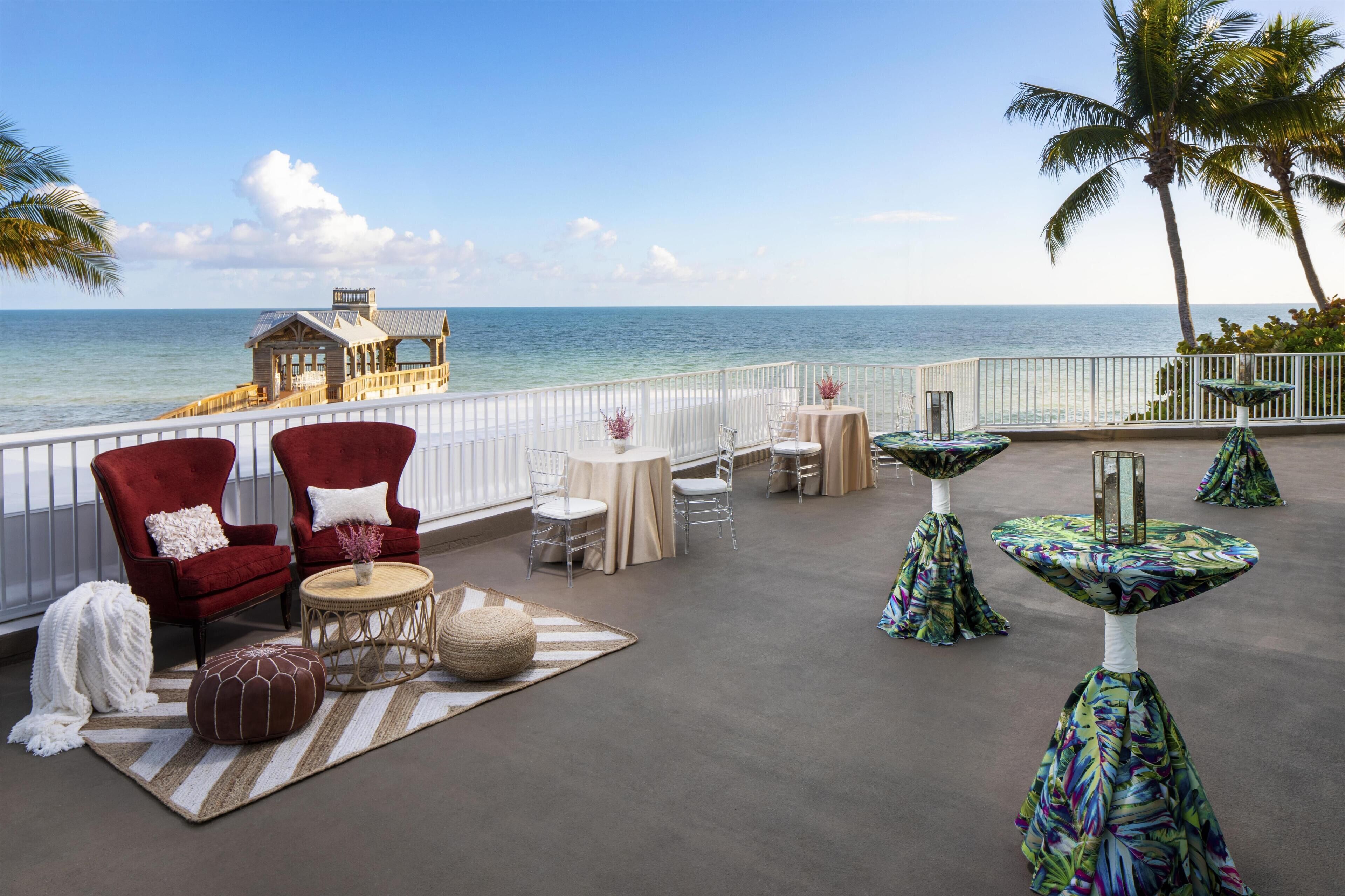 A coastal terrace with red and white chairs, round tables, and a view of the ocean.