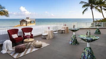 A coastal terrace with red and white chairs, round tables, and a view of the ocean.