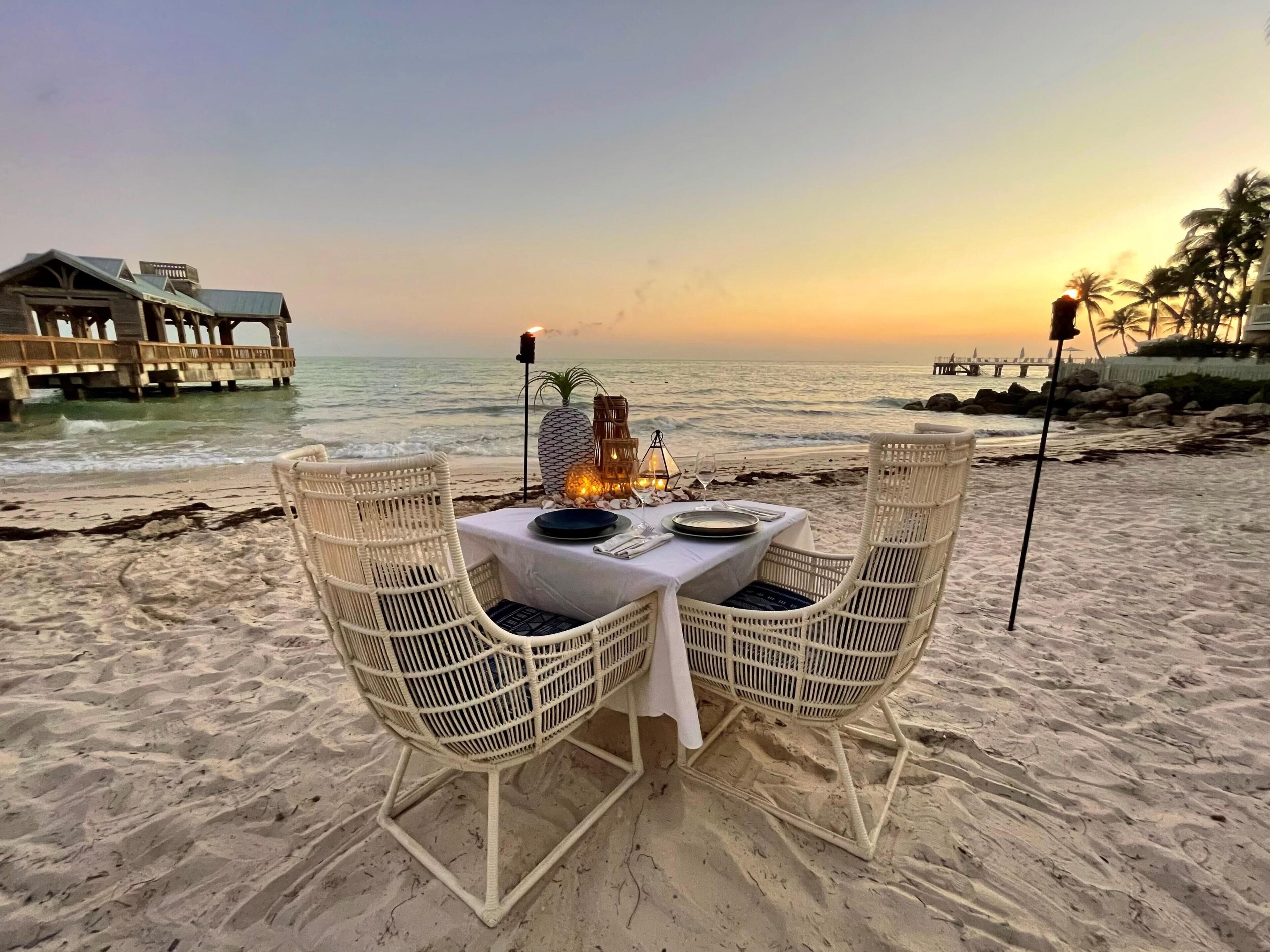 Un ambiente para comer frente a la playa con sillas de mimbre, una mesa para dos personas y vista al océano y un muelle al fondo.