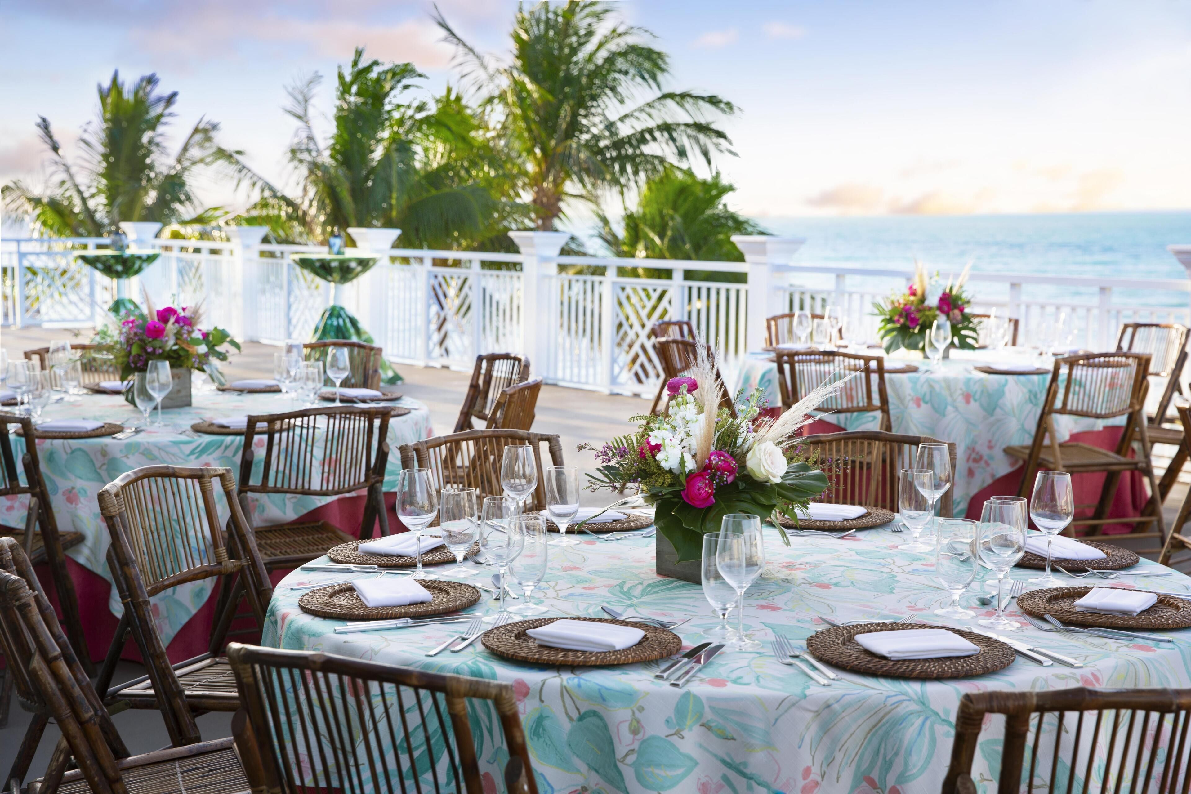 A seaside dining area with round tables, wicker chairs, and floral centerpieces.
