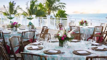 A seaside dining area with round tables, wicker chairs, and floral centerpieces.