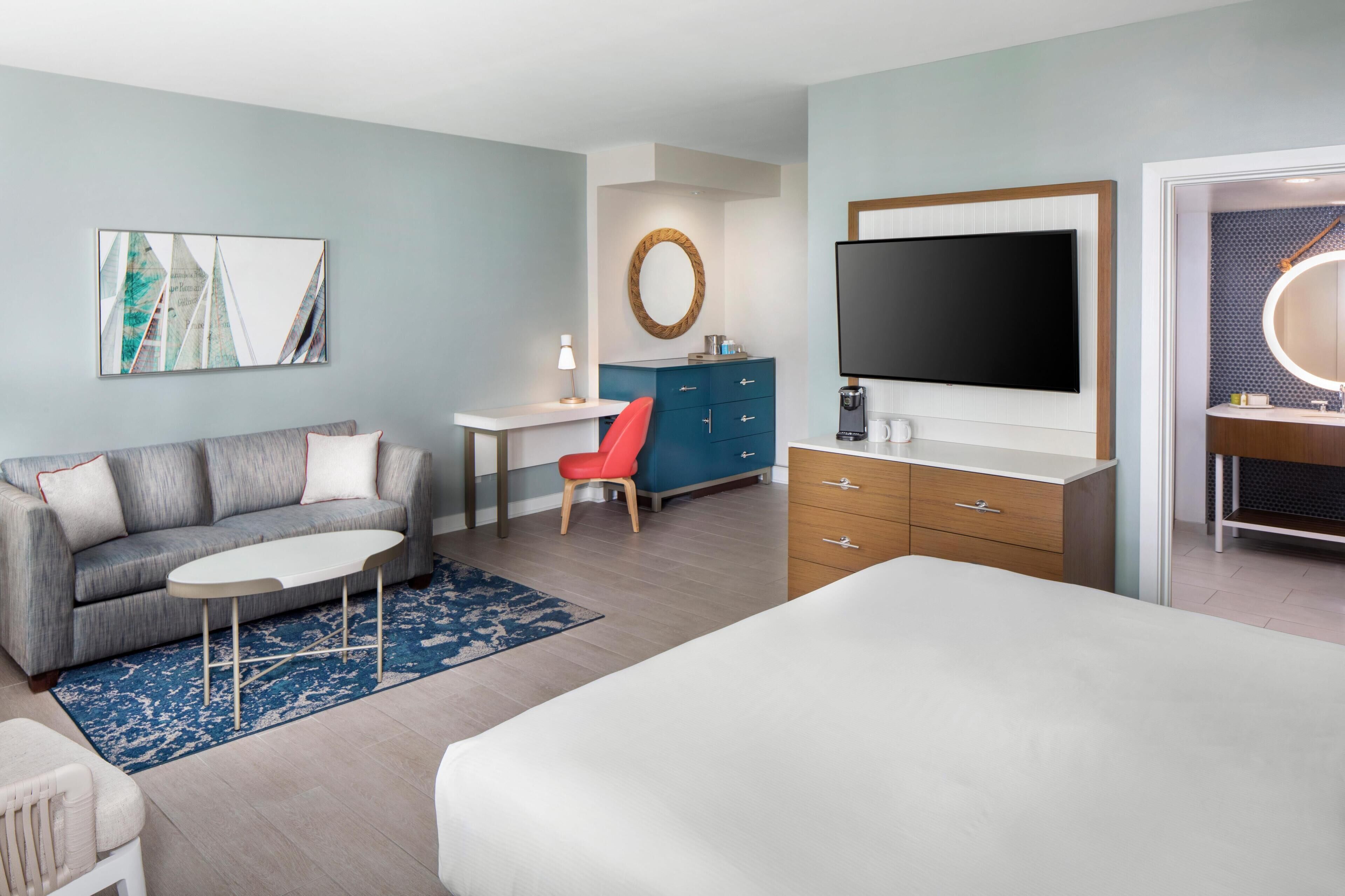 A modern hotel room with a grey sofa, a red chair, a white bed, a TV, a wooden cabinet, and a desk with a lamp.