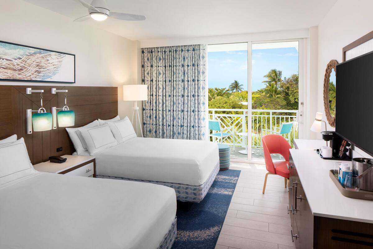 Hotelkamer met twee bedden, een bureau, een televisie en een balkon met uitzicht op palmbomen.
