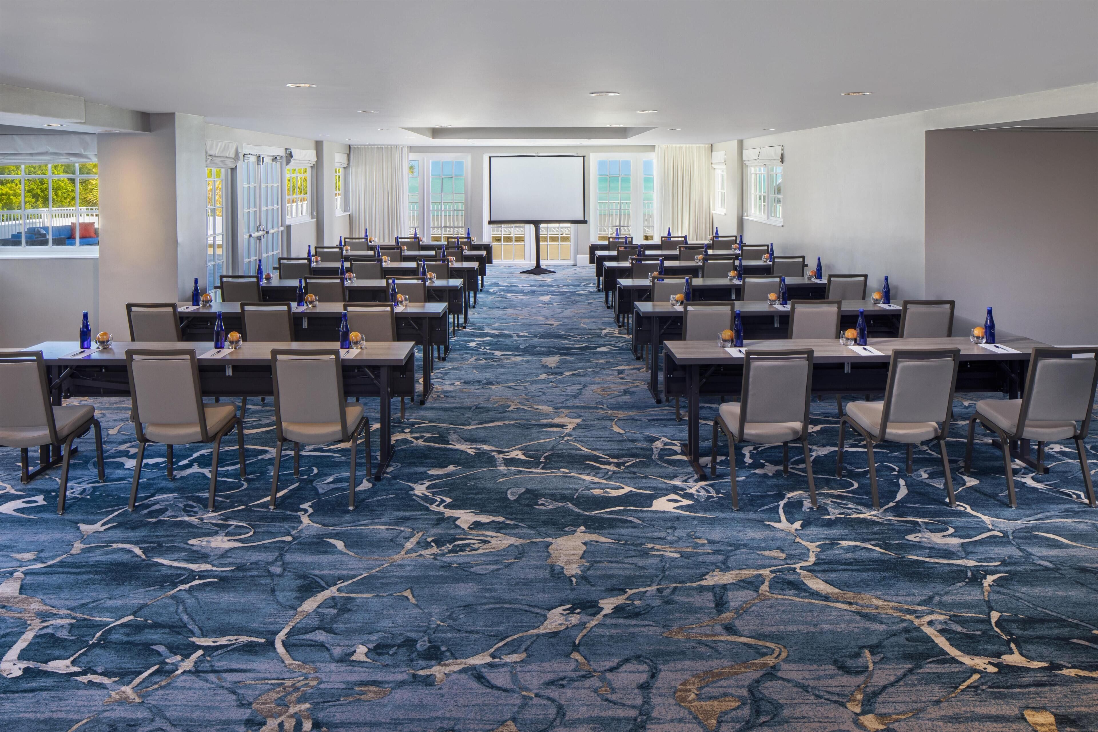 A conference room with rows of tables and chairs, a large screen at the front, and a carpeted floor.