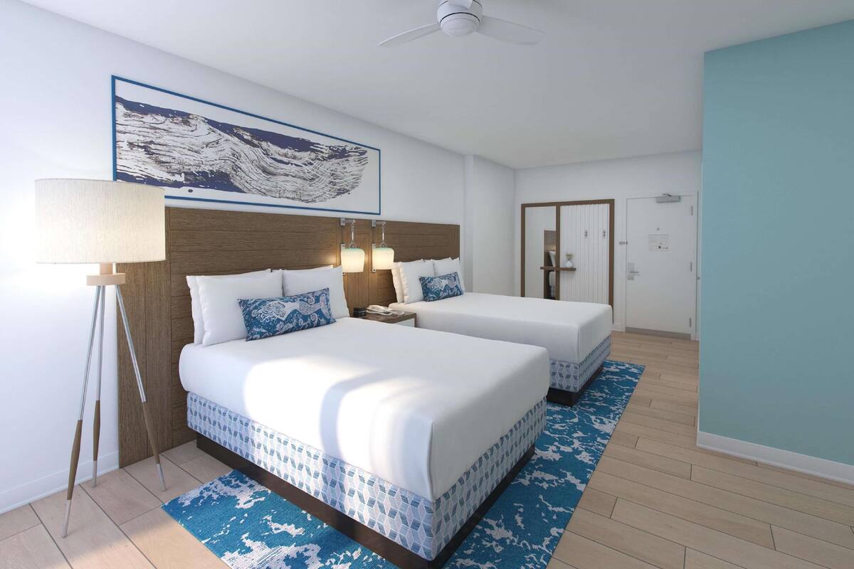 Een hotelkamer met twee bedden, een groot schilderij, een plafondventilator en een blauw kamerkleed.