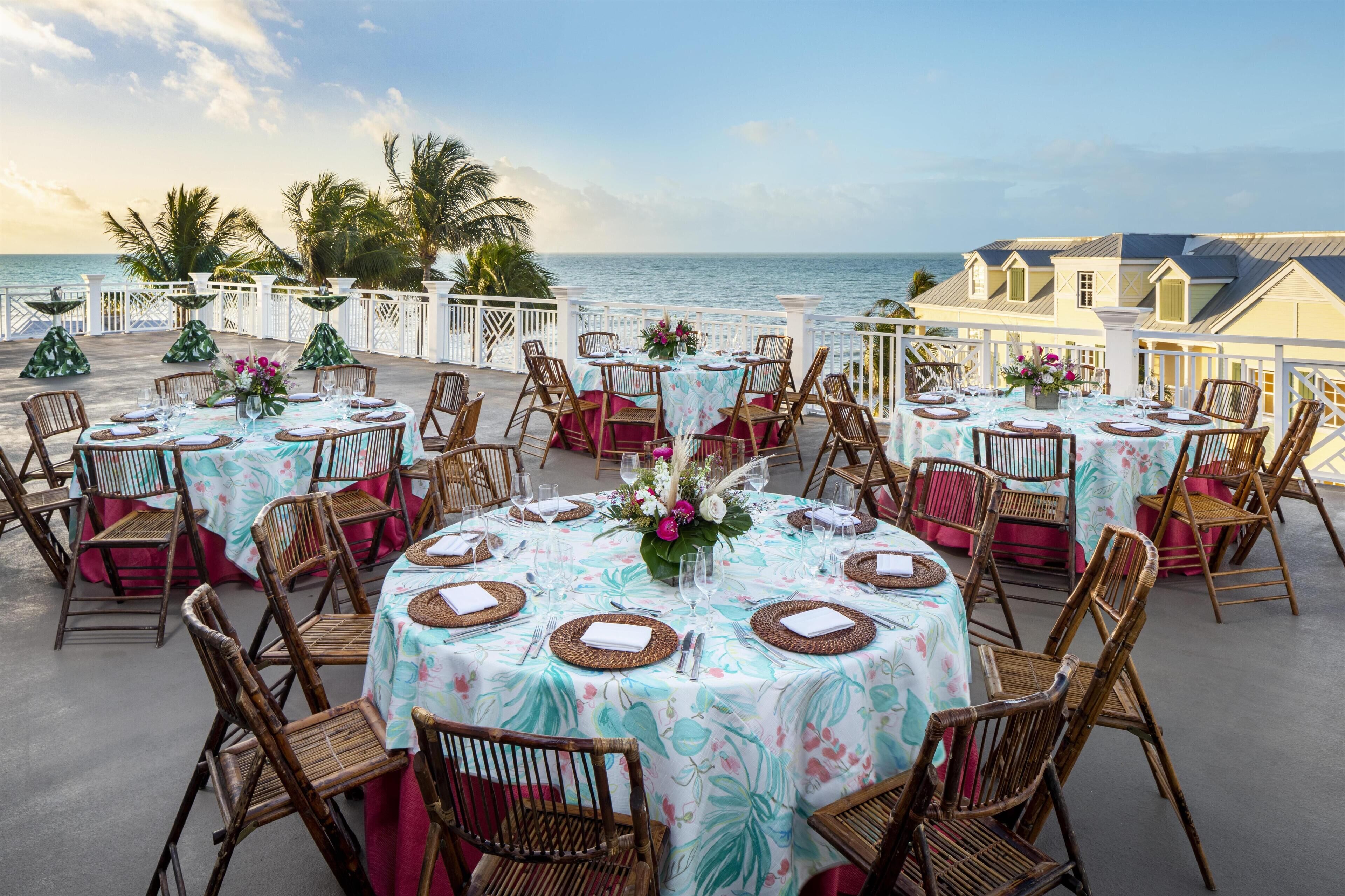 A seaside dining area with round tables set for guests, featuring wicker chairs and floral tablecloths.