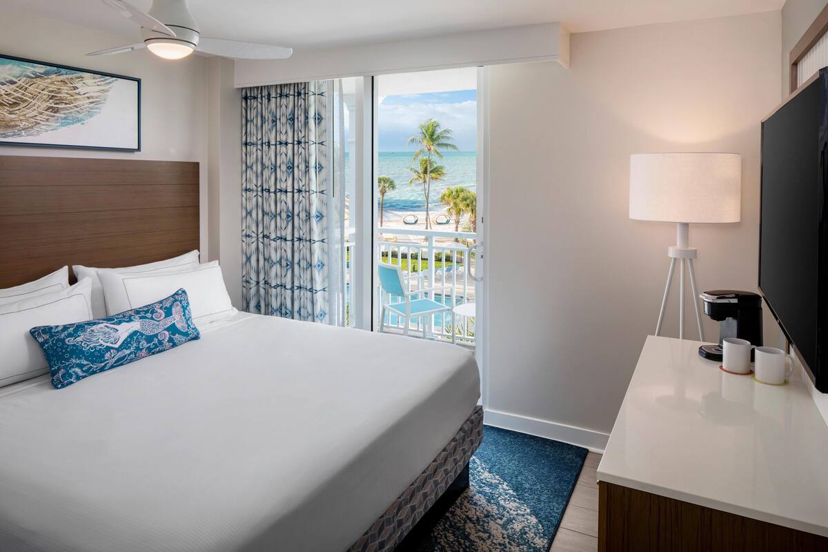 Een hotelkamer met een bed, een bureau, een televisie en een balkon met uitzicht op de oceaan.