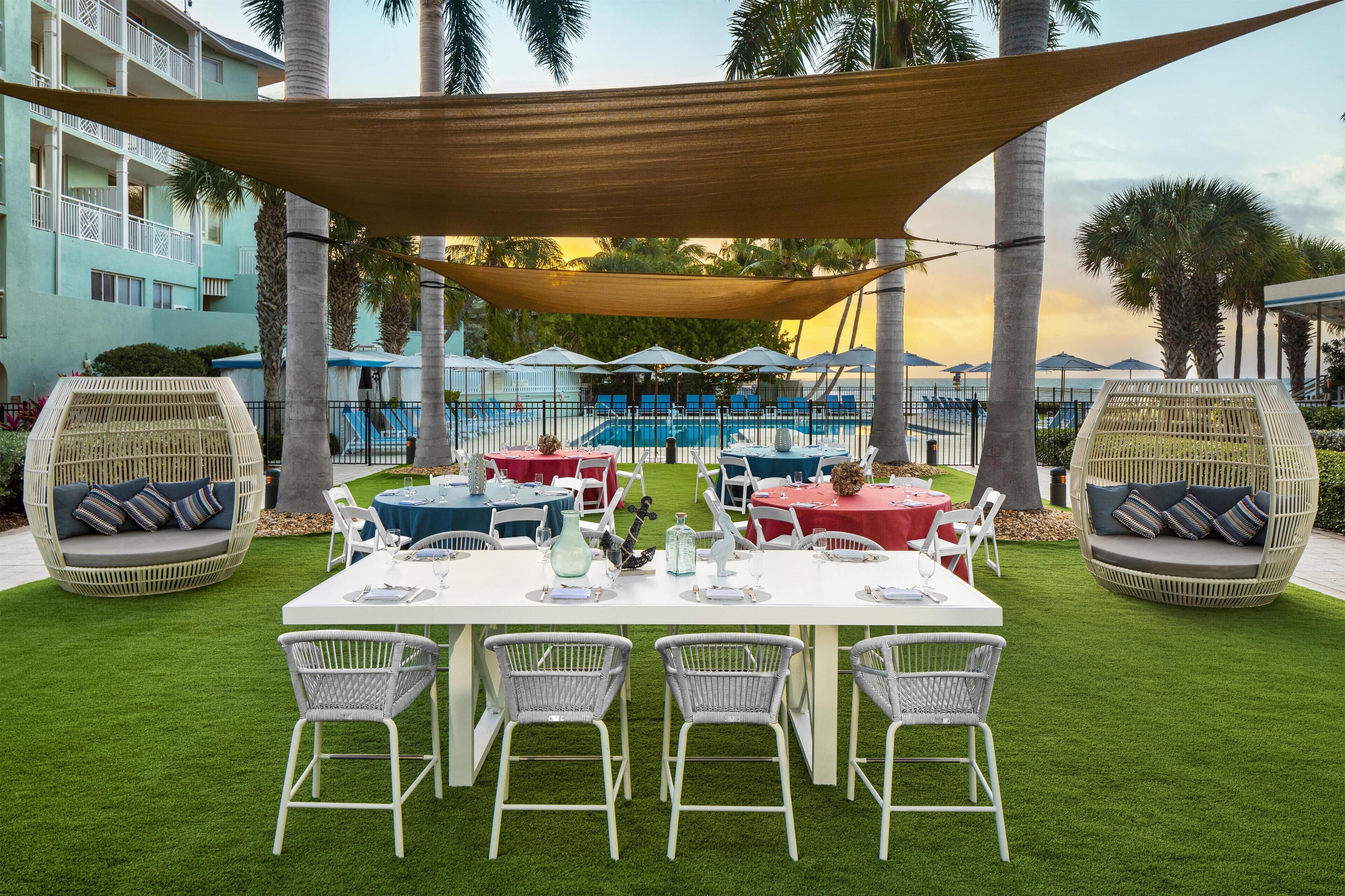 An outdoor dining area with tables and chairs, surrounded by palm trees and a pool.