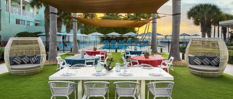 An outdoor dining area with tables and chairs, surrounded by palm trees and a pool.