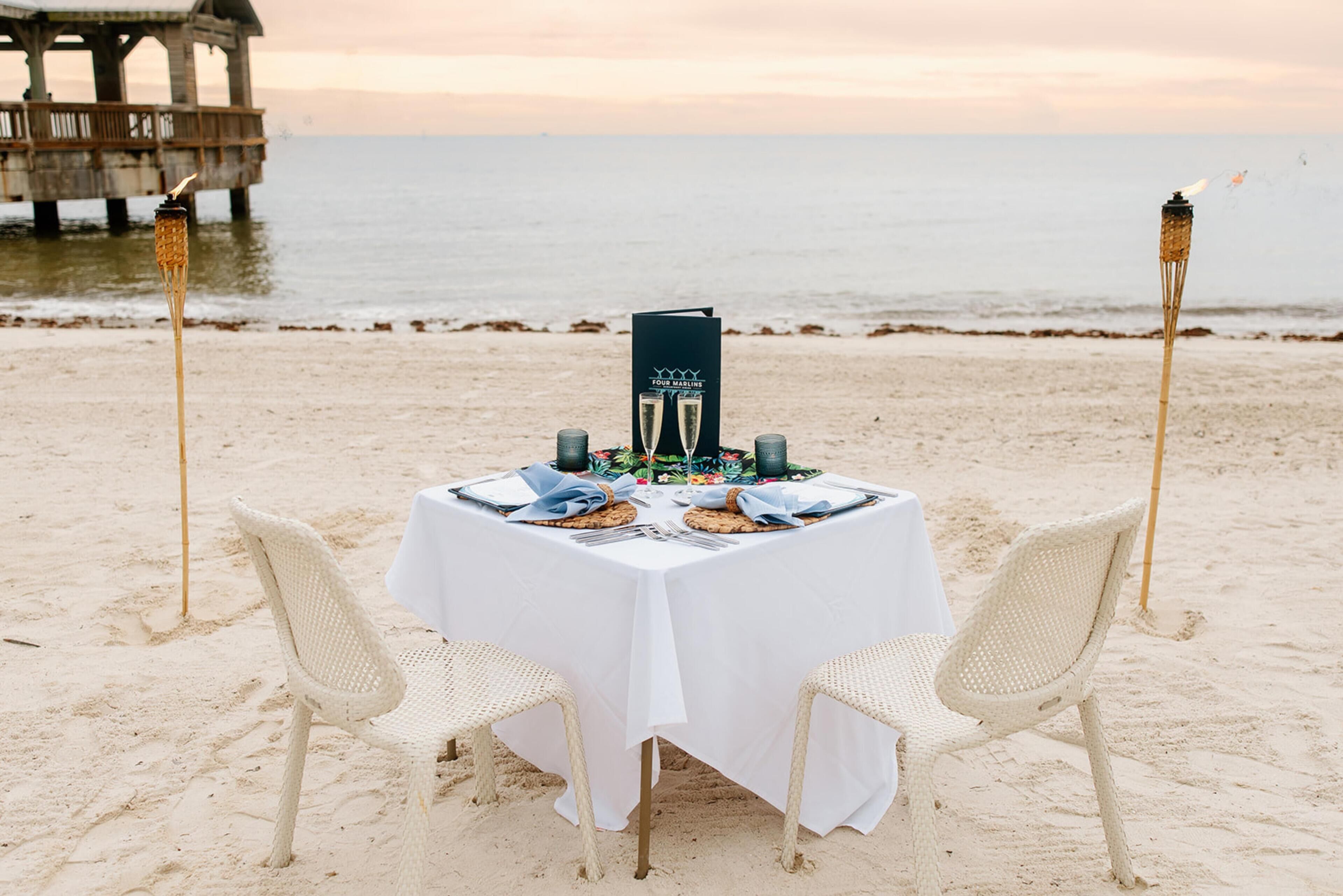 A table set for two with a white tablecloth, two chairs, and a menu stand on a beach with a pier in the background.
