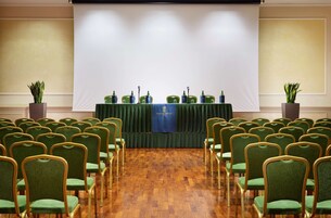 A conference room with rows of green chairs, a stage with a banner, and potted plants.
