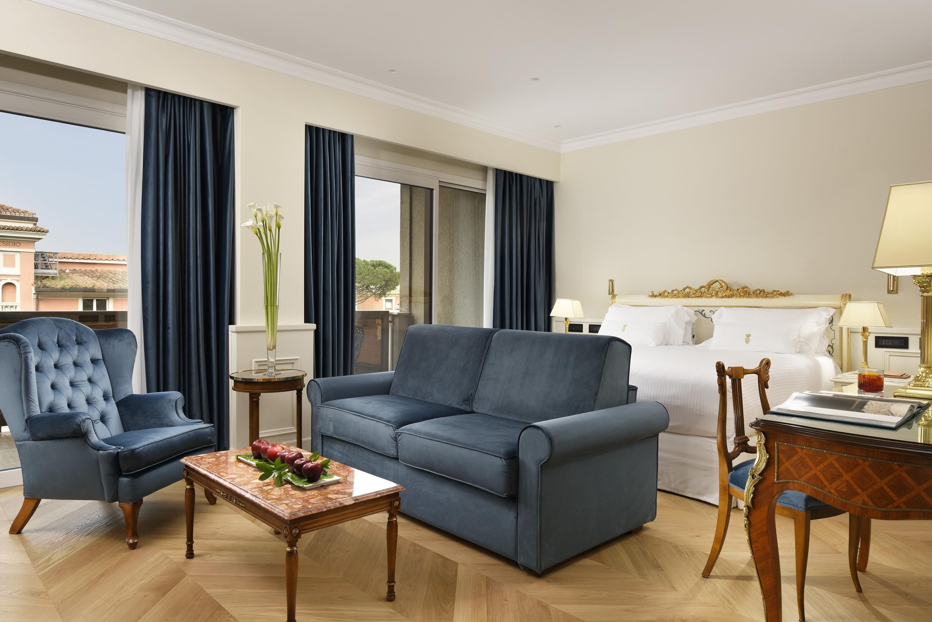 Una habitación de hotel con un sofá azul, una silla de madera, una mesita con un florero y una cama con ropa de cama blanca, y vistas a edificios.