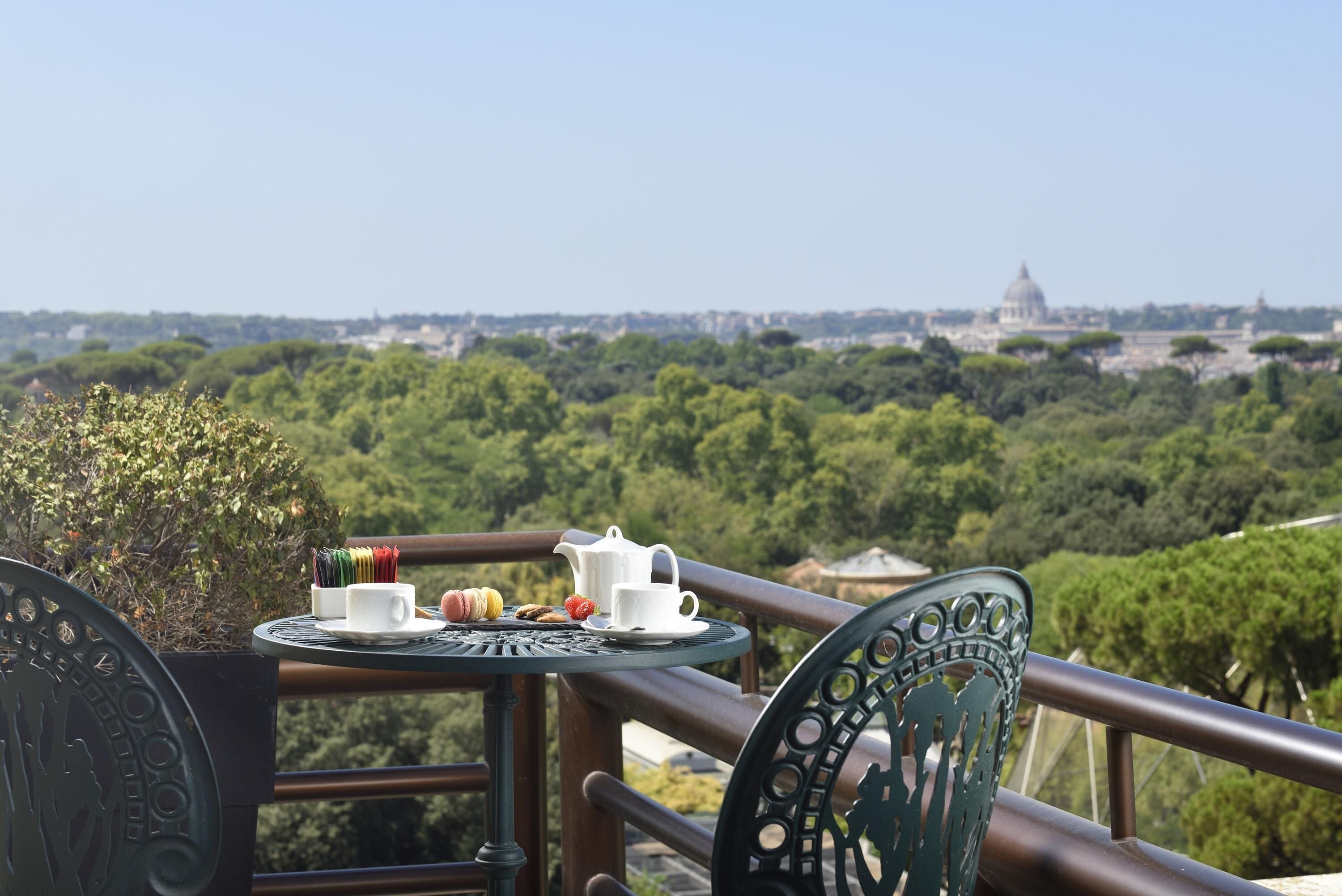 A balcony with a table set for tea, overlooking a lush green landscape and a distant building.