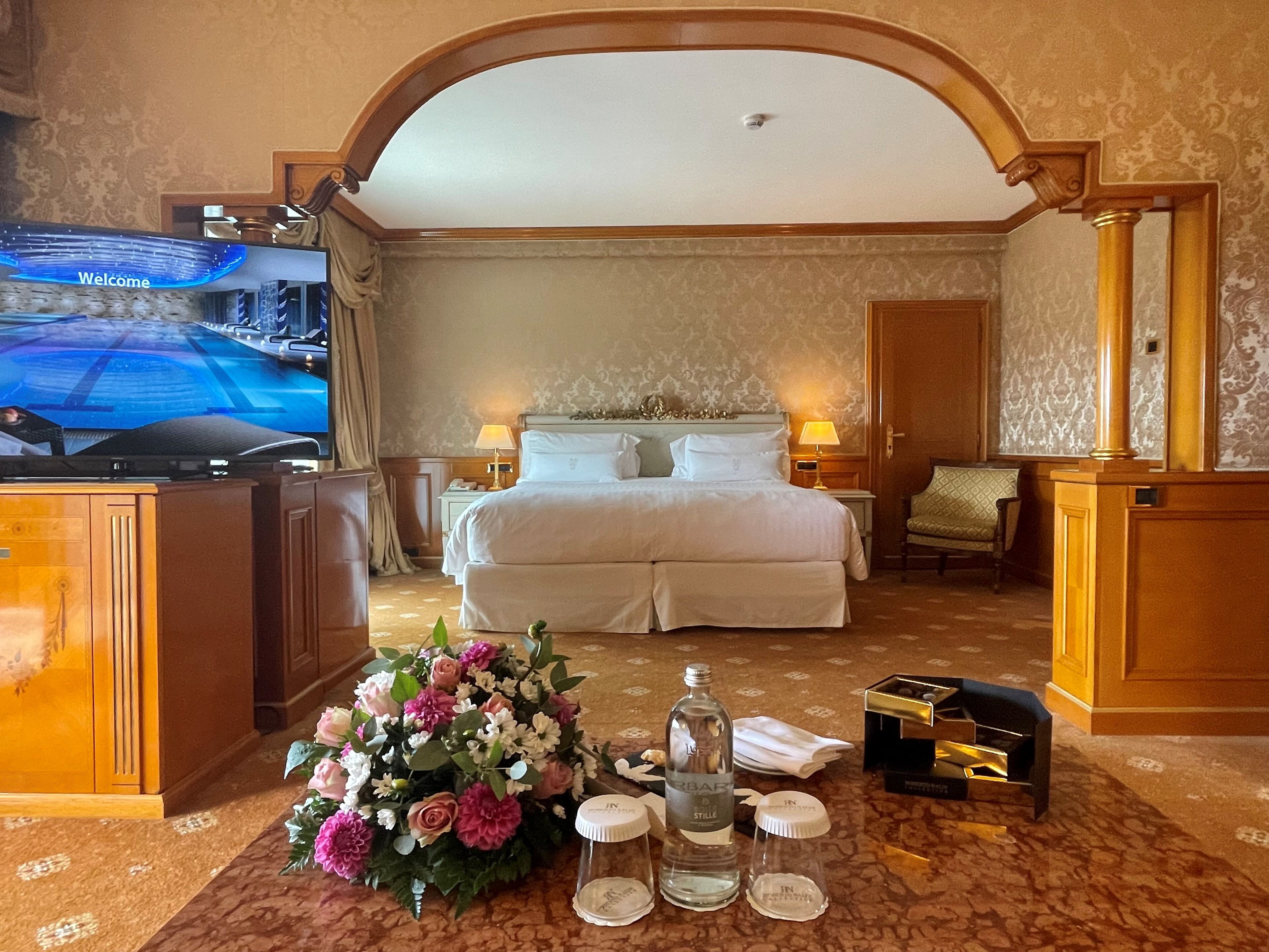 A hotel room with a large bed, a television displaying a welcome message, a floral arrangement, and a minibar.