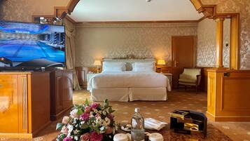 A hotel room with a large bed, a television displaying a welcome message, a floral arrangement, and a minibar.