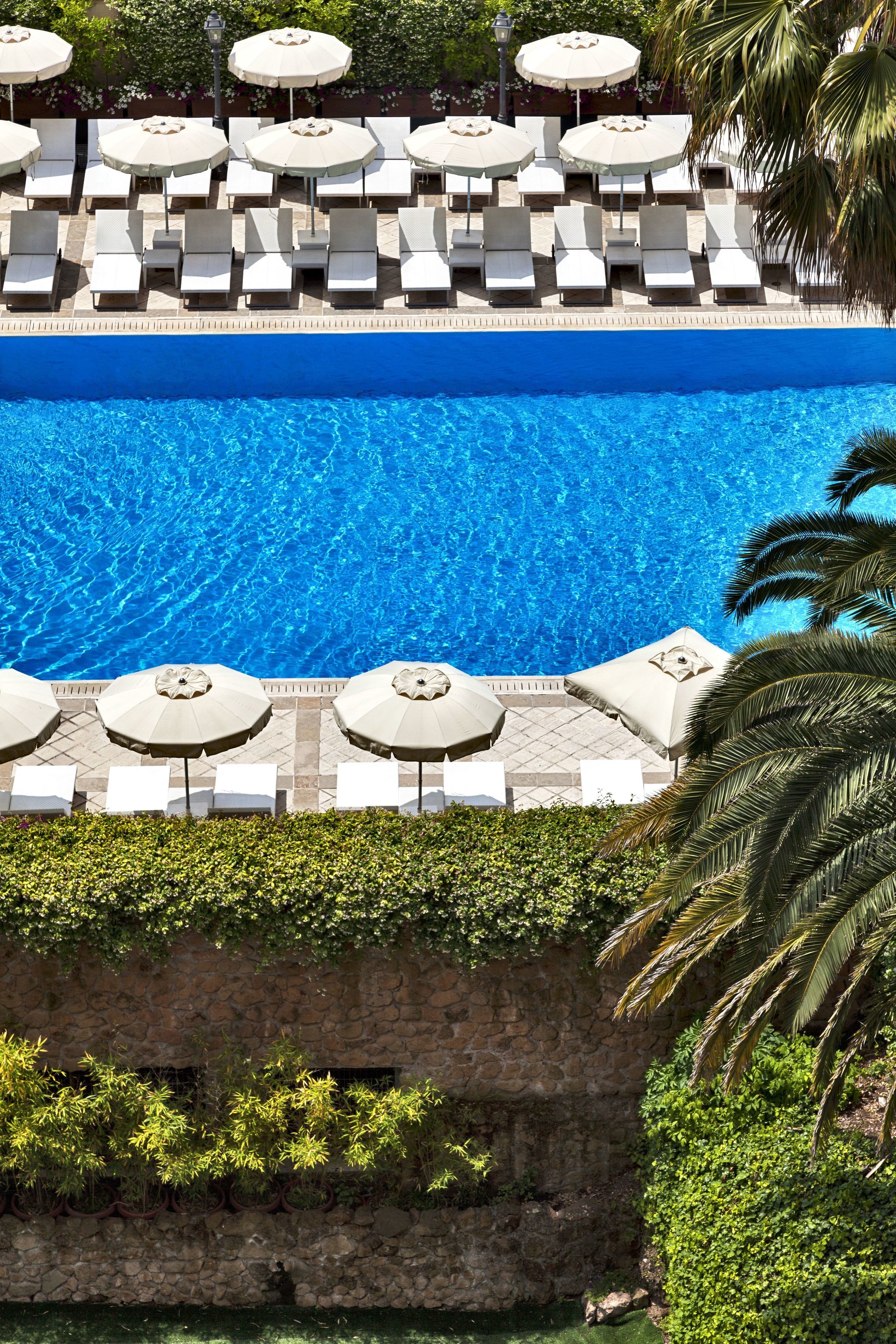Un espace piscine agrémenté de chaises longues et de parasols, entouré de verdure et délimité par un mur de pierre.