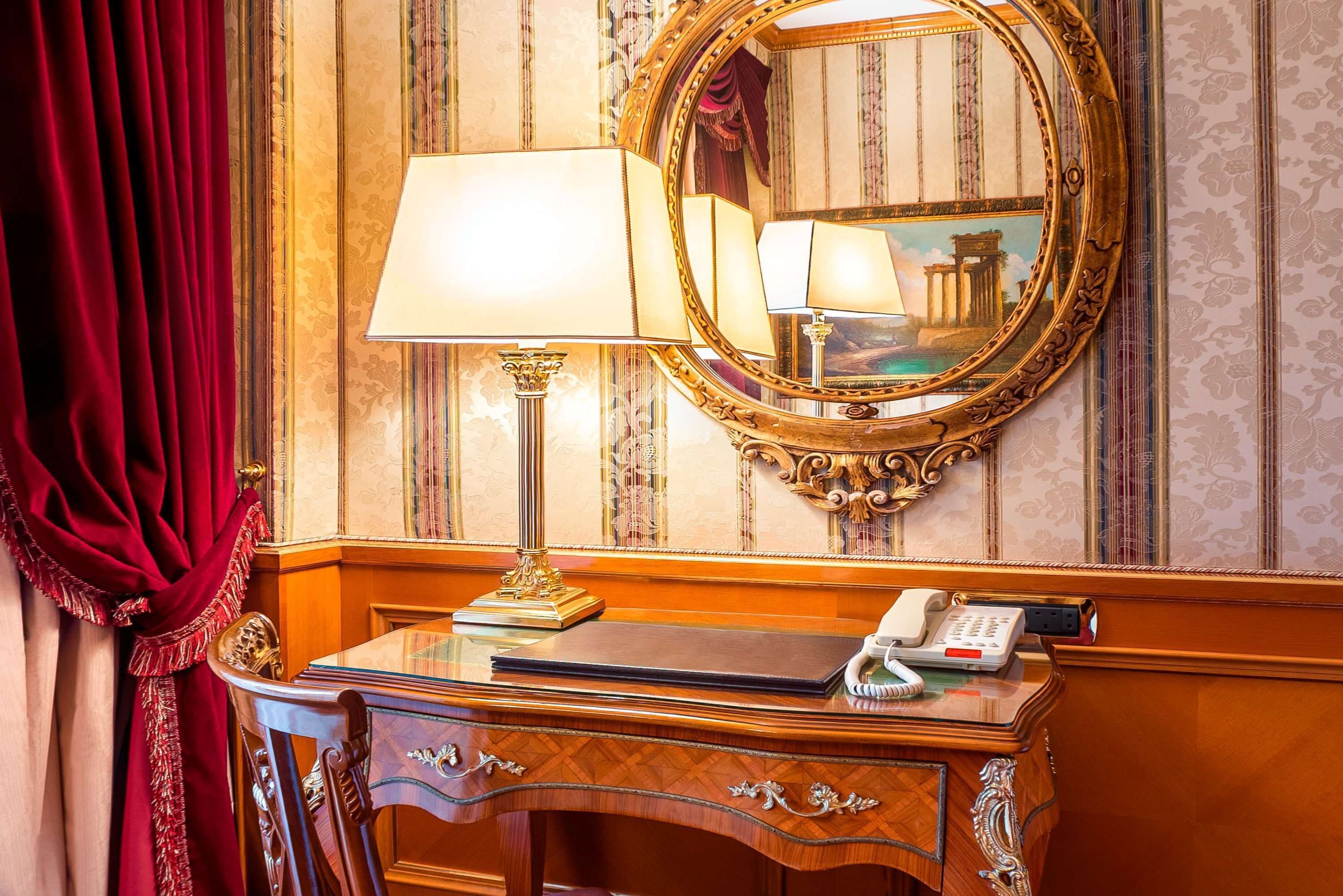 A wooden desk with a lamp, a telephone, and a mirror with a decorative frame.