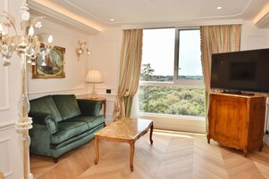 A living room with a green sofa, a wooden coffee table, a TV on a wooden cabinet, and a large window with curtains.