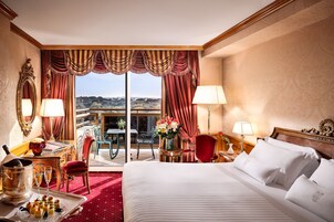 A hotel room with a large bed, two bedside tables, a desk with a chair, a dining area with a table and chairs, and a view of the outdoors.