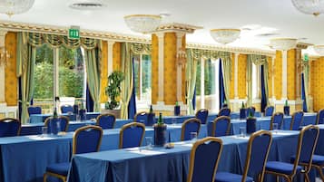 Una sala da banchetto con file di tavoli, tovaglie blu e finestre con cornice dorata.