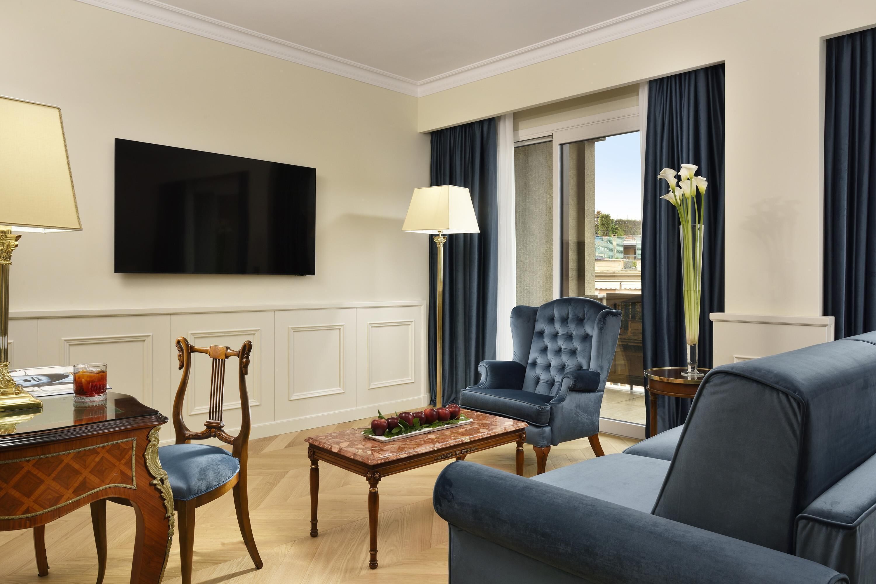 Quarto de hotel com uma televisão de tela grande fixada na parede, uma mesa de centro de madeira com frutas, uma poltrona azul, um sofá e vista para o exterior por uma porta de vidro deslizante.
