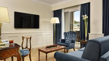 Quarto de hotel com uma televisão de tela grande fixada na parede, uma mesa de centro de madeira com frutas, uma poltrona azul, um sofá e vista para o exterior por uma porta de vidro deslizante.