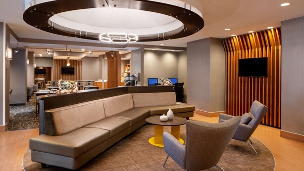 A modern hotel lobby with a curved sofa, a round coffee table, and a seating area with armchairs. There is a reception desk with a flat-screen TV and a kitchen area in the background. - Sonesta Select Nashville Airport Suites (Nashville)