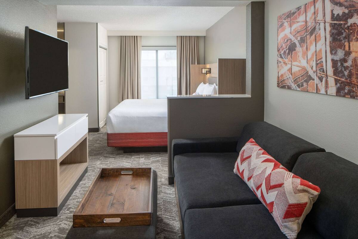 A hotel room with a flat-screen TV, a bed, a sofa, a wooden side table, and a large abstract painting on the wall.